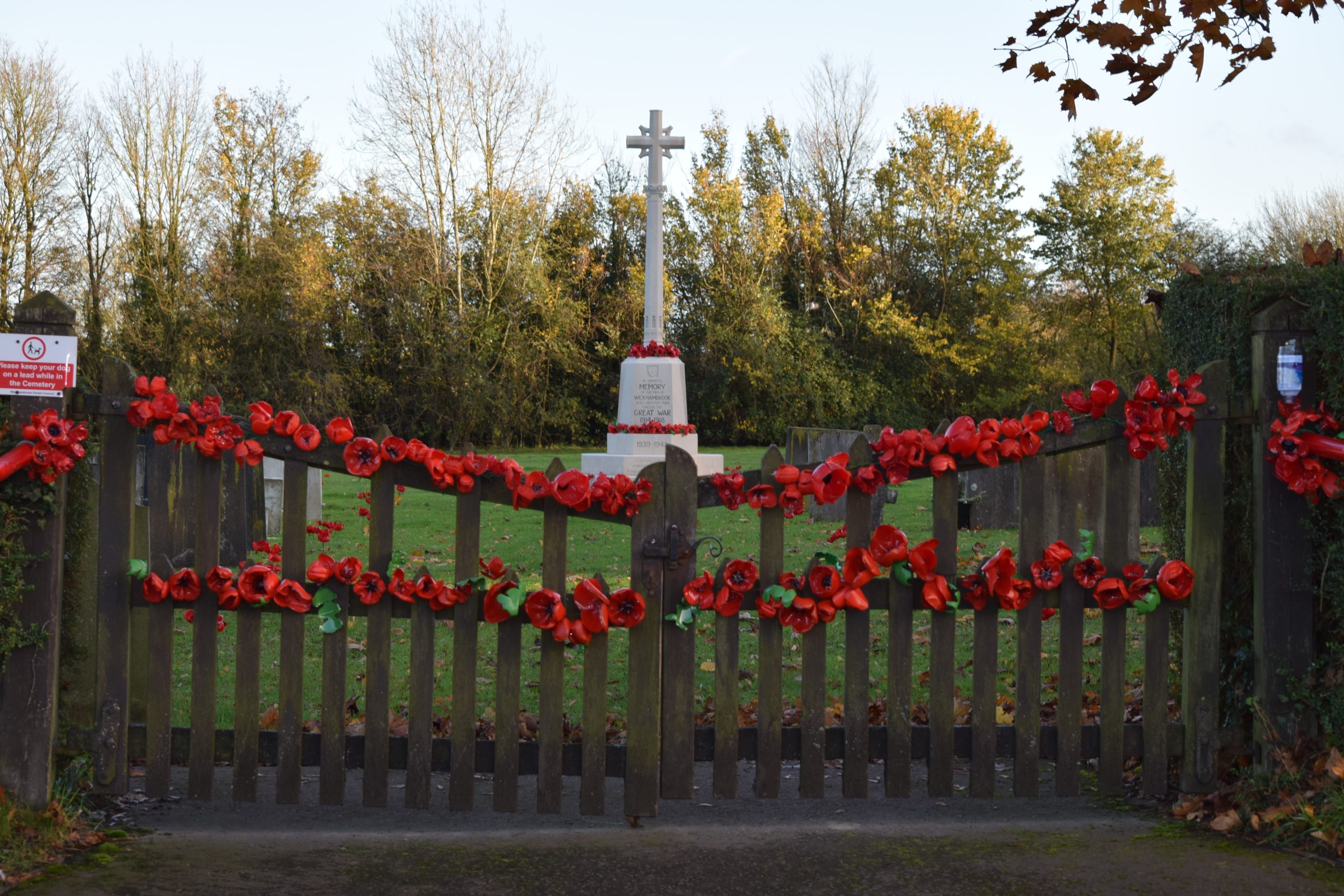 Photograph showing a remembrance memorial beside a wooden gate, dedicated to fallen service personnel. Red poppy wreaths line gate, with a white pillar and cross at centre, small plaques, and autumn trees forming background.
