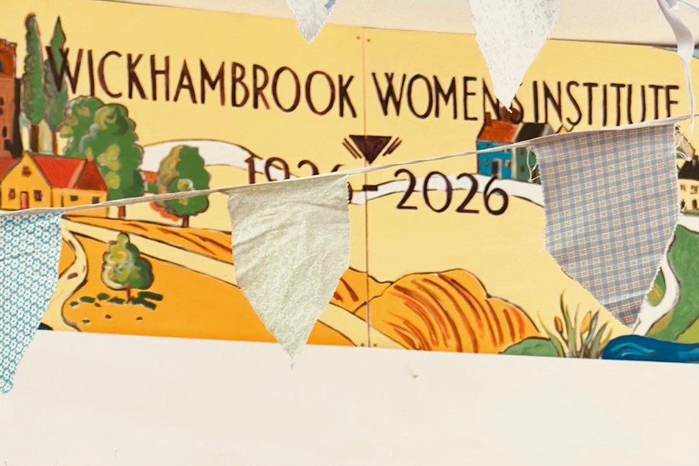 Photograph of part of a colourful mural and fabric bunting. Mural displays "Wickhambrook Women’s Institute 1926-2026," indicating a centenary celebration, with a village scene and decorative elements in warm tones.