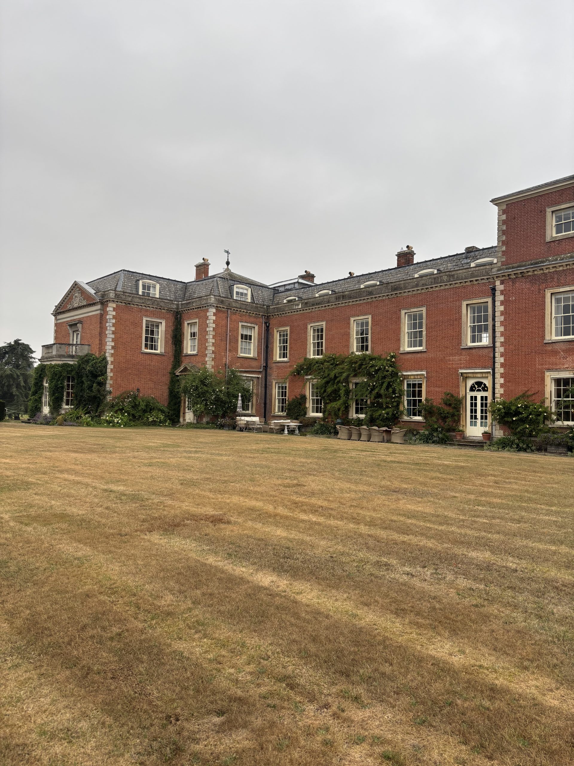 Photograph of a large red-brick Georgian-style building with multiple chimneys and white-framed windows, set against an overcast sky. Foreground features a wide, dry grass lawn with visible patchy brown areas, indicating lack of recent rainfall.