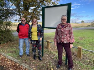 A group of people standing next to a noticeboard with trees and farmland in the background