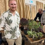 Photograph showing smiling man in botanical-print shirt beside a table of potted plants, likely at an indoor gardening or plant sale event. Older visitors examine assorted leafy plants and flowers, with patterned curtains, folding tables and illuminated emergency-exit sign in background.