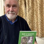 Photograph of an older man with white beard holding a green booklet titled “Bury St Edmunds: A compilation”. Booklet features historical photographs and text, with patterned curtains and indoor setting behind him.